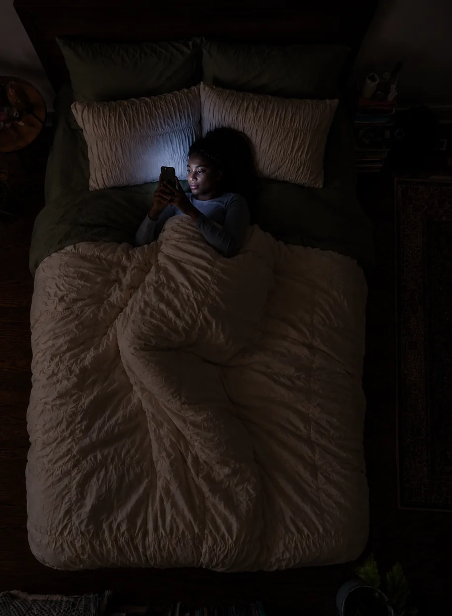 A woman scrolling on her phone in bed late at night, her face lit by the screen